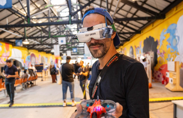 Primeiro plano de um homem sorrindo, usando óculos de realidade virtual brancos e um boné azul virado para trás. Ele segura em primeiro plano um pequeno drone com luzes acesas. O homem está vestido com uma camiseta de manga longa preta e uma fita pendurada no pescoço. O cenário ao fundo é o interior de um grande galpão, com teto de estrutura metálica, paredes decoradas com grafites amarelos e outras pessoas circulando pelo evento.