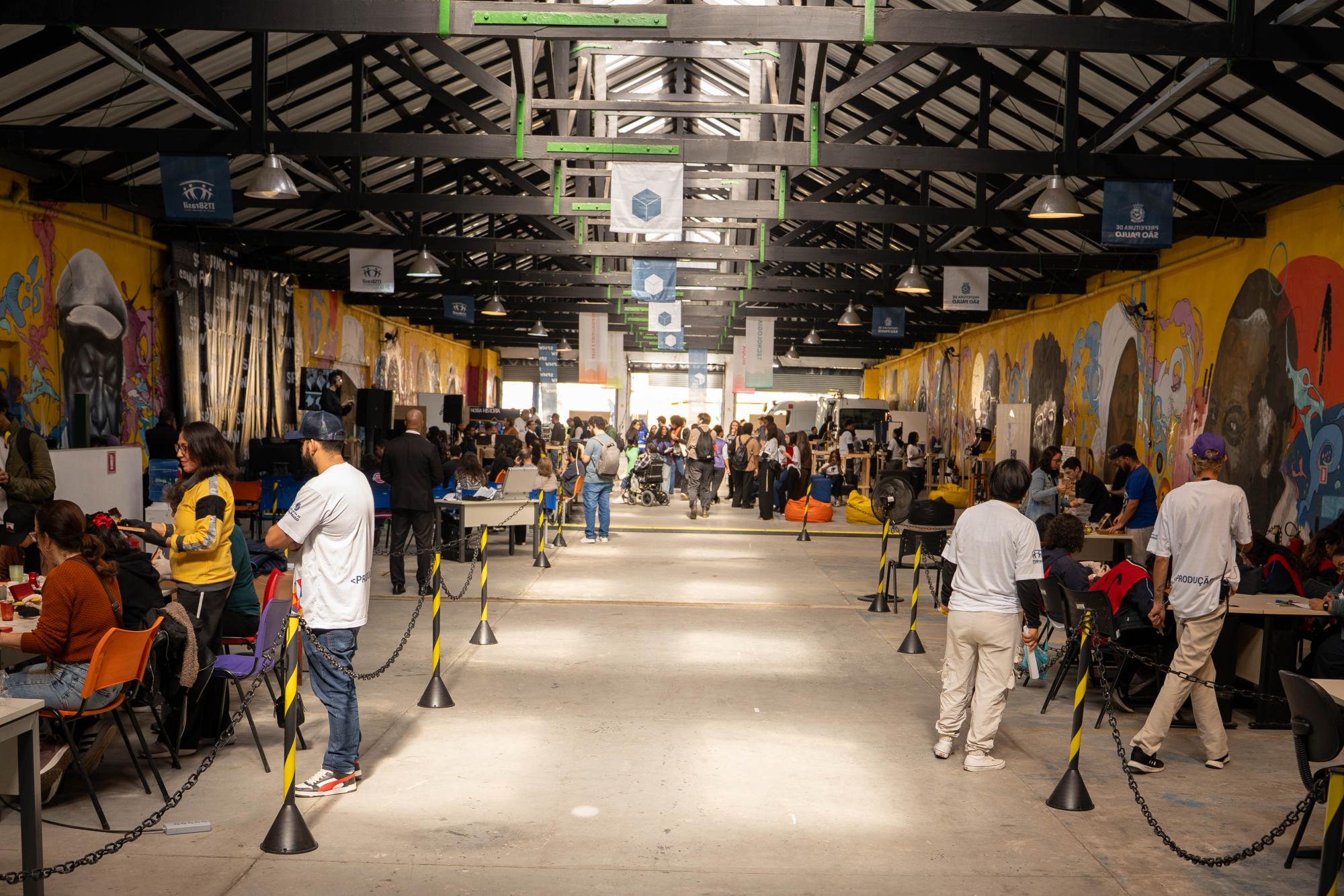 coloridos. O ambiente mostra o público circulando livremente pelo local e interagindo em mesas de trabalho. No teto de estrutura exposta, diversas bandeiras e faixas verticais coloridas estão penduradas ao longo do espaço.
